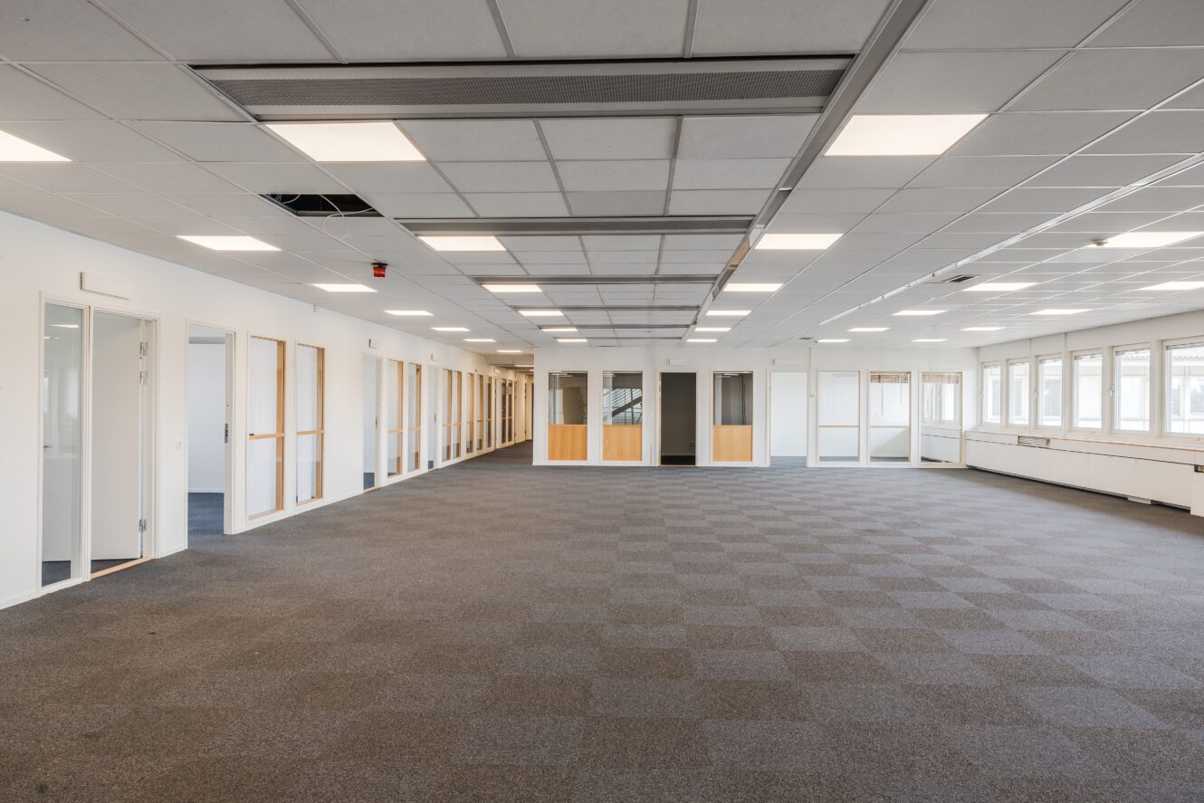 Stort, tomt kontorsutrymme med grå heltäckningsmatta, vita väggar, flera kontorsdörrar med glaspaneler och ett gallertak med lysrör.