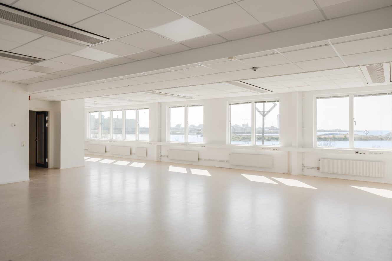 Tomt, rymligt rum med vita väggar, stora fönster som släpper in dagsljus, beige golv och utsikt över byggnader och vatten utanför. Taket har infällda lampor och kakel.
