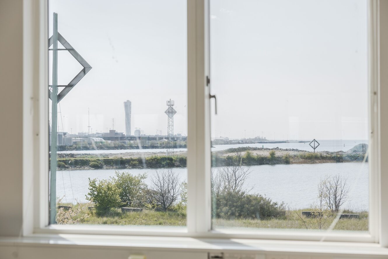 Från ett stort fönster har man utsikt över ett vattendrag, en gräsyta och avlägsna industribyggnader med torn och metallkonstruktioner under en disig himmel.