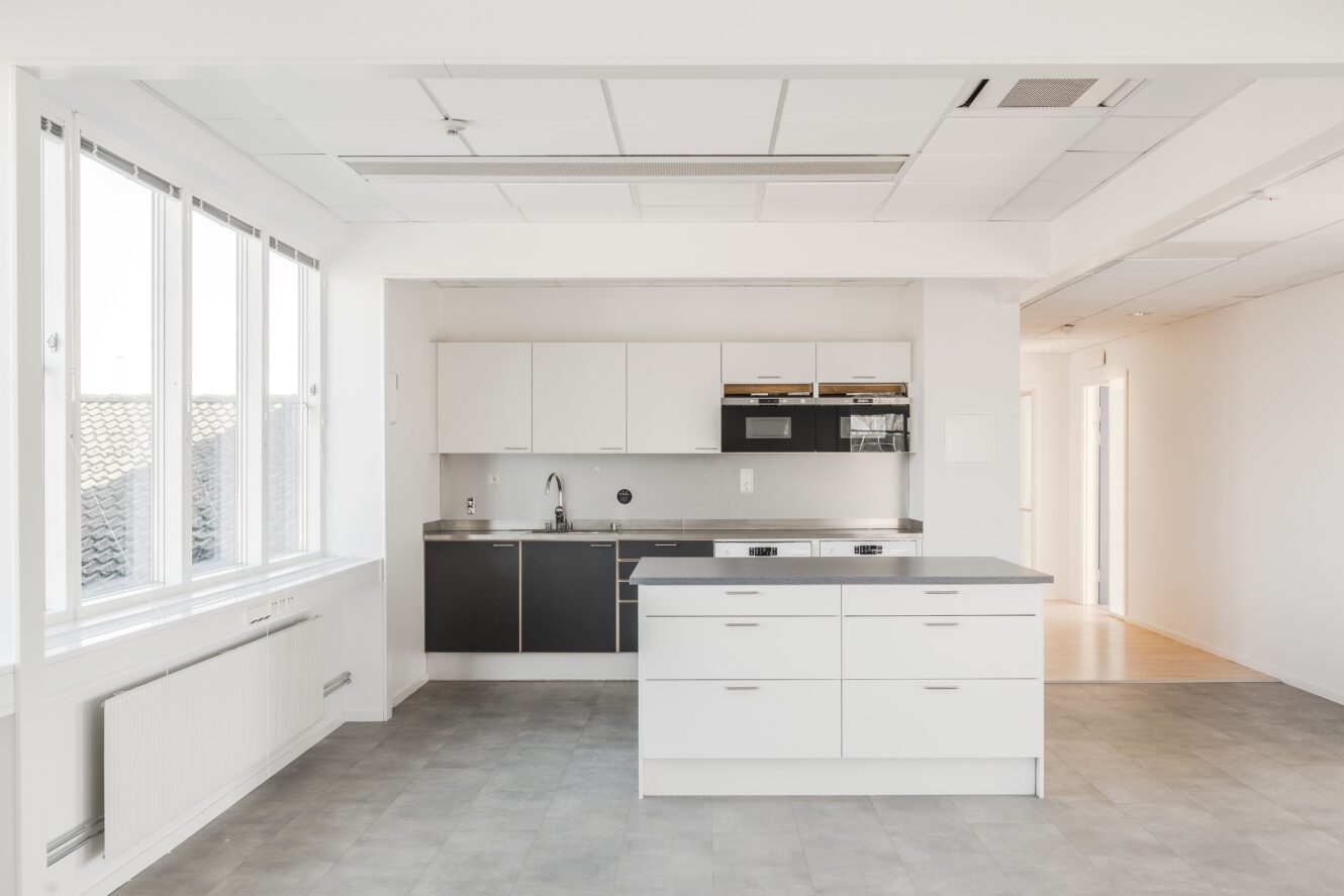 Ett modernt, minimalistiskt kök med vita och svarta skåp, en central köksö, vitvaror i rostfritt stål och stora fönster som släpper in naturligt ljus.