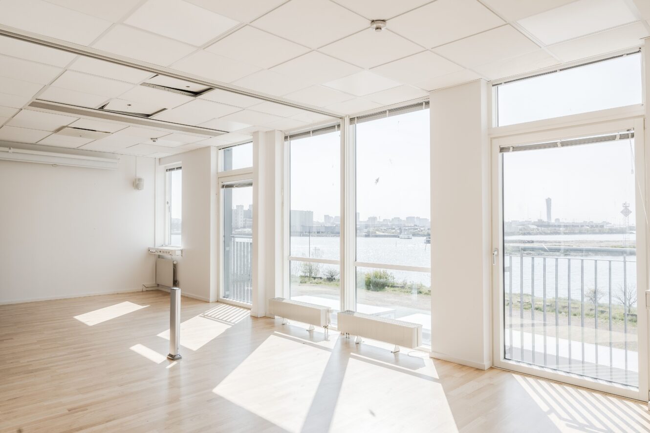 Ljust, tomt rum med stora fönster, ljust trägolv, vita väggar och utsikt över en flod och en stadsbild utanför. Takplattor och radiatorer är synliga.