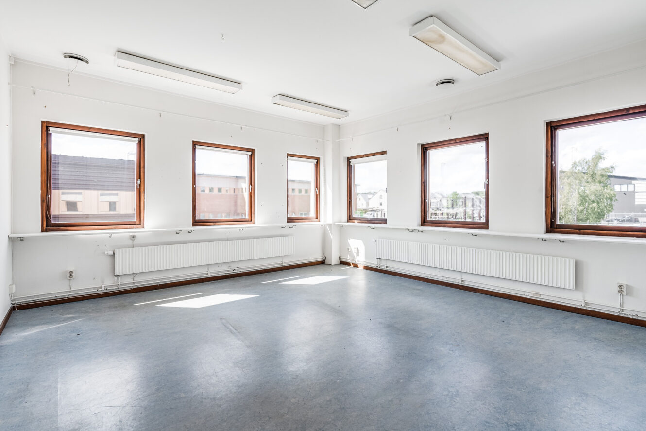 Tomt, ljust rum med fem stora fönster, vita väggar, blågrått golv, taklampor och radiatorer under fönstren. Dagsljus kommer in genom fönstren.