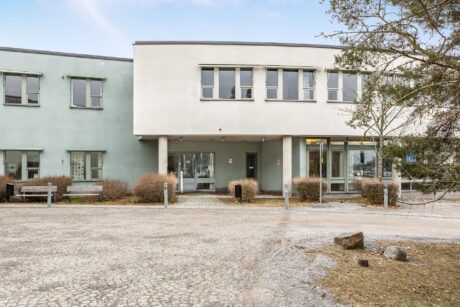 En modern tvåvåningsbyggnad med ljusgröna och vita ytterväggar, flera fönster, en entré med pelare, bänkar och lövfällande buskar framför.