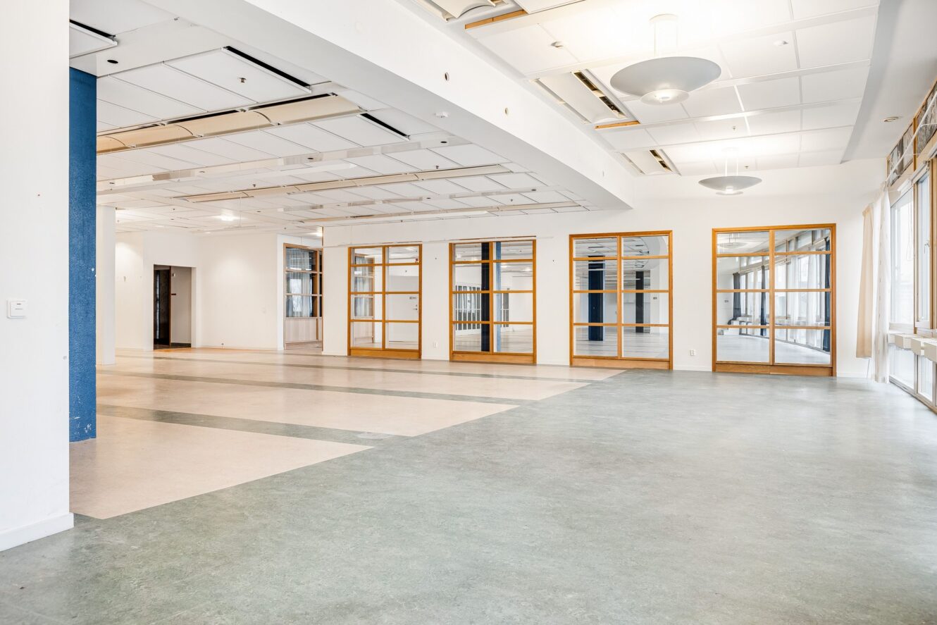 Rymligt, tomt rum med stora fönster, trädörrar med glaspaneler och vita takplattor med moderna armaturer. Naturligt ljus kommer in från höger sida.