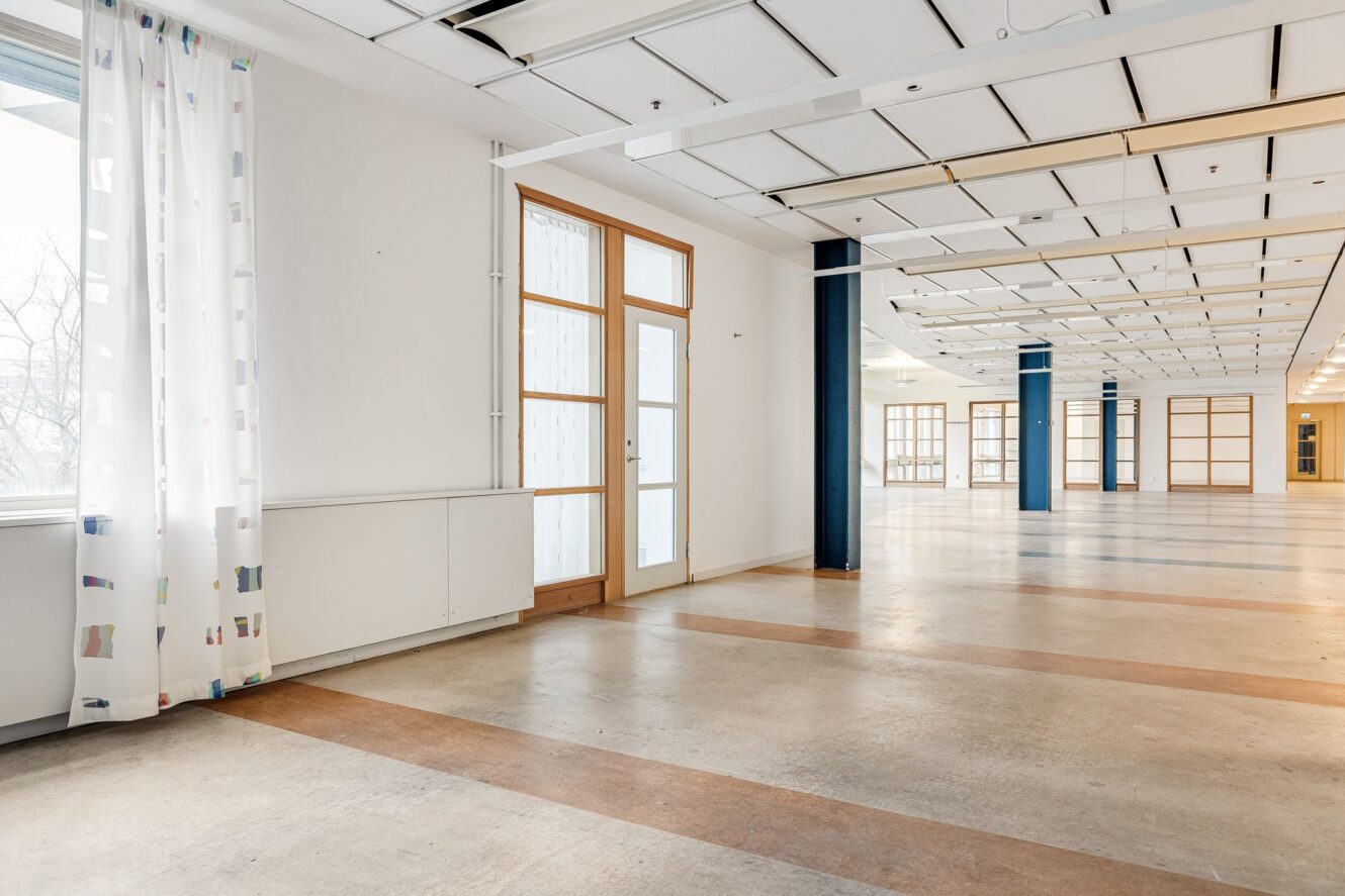 Rymligt, tomt rum med stora fönster, glasdörrar, vita väggar, synliga takpaneler och mönstrade gardiner, med naturligt ljusinsläpp och ett polerat betonggolv.