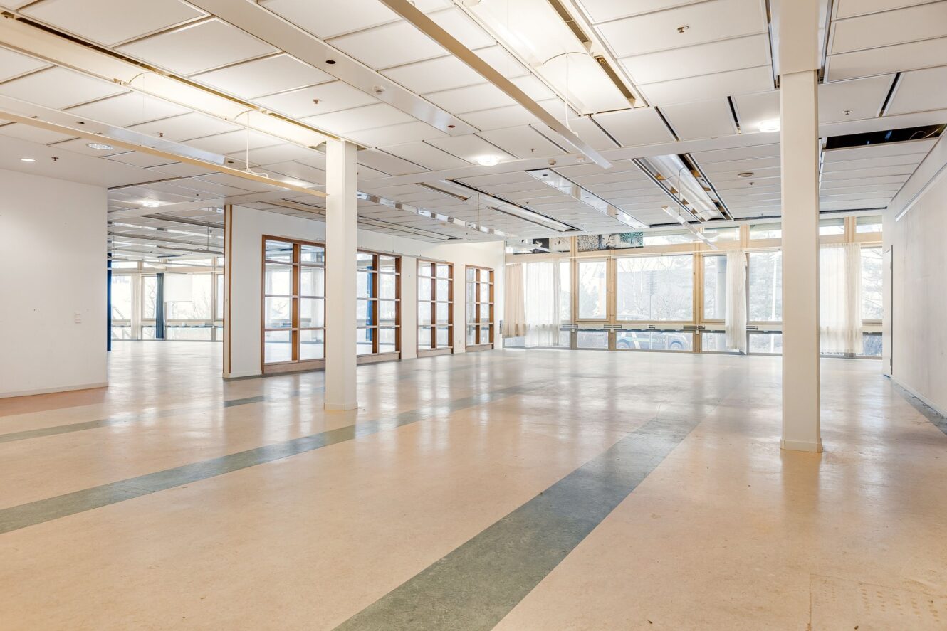 Rymligt, tomt rum med stora fönster, vita väggar, taklampor och gröna golvremsor. Naturligt ljus fyller utrymmet genom glasdörrar och fönster.