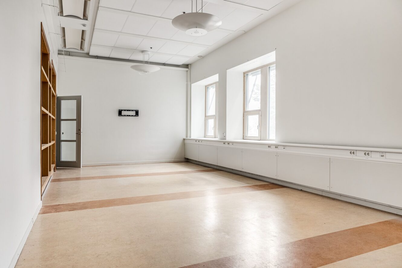 Tomt rum med vita väggar, beige golv med bruna ränder, två stora fönster, taklampor, trähyllor till vänster och en stängd dörr längst bak.