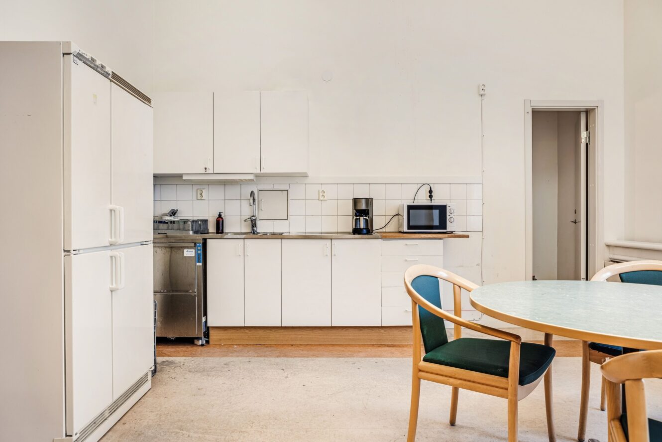 Ett enkelt kök med vita skåp, kylskåp, mikrovågsugn, kaffebryggare, diskho och ett runt bord med fyra stolar.