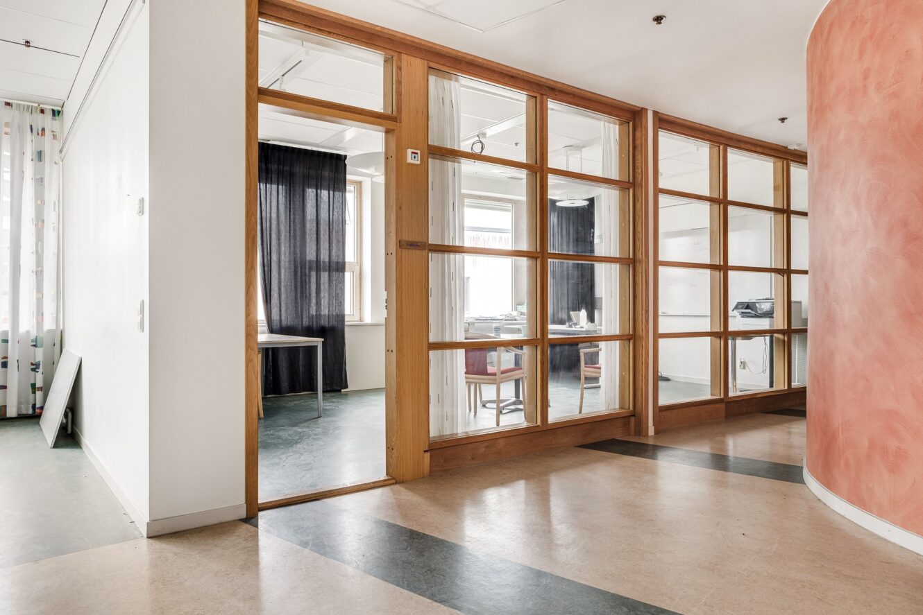 En hall med kontorsdörrar av trä med glaspaneler, beige och grönt golv och en böjd vägg målad i en rödaktig strukturfärg.