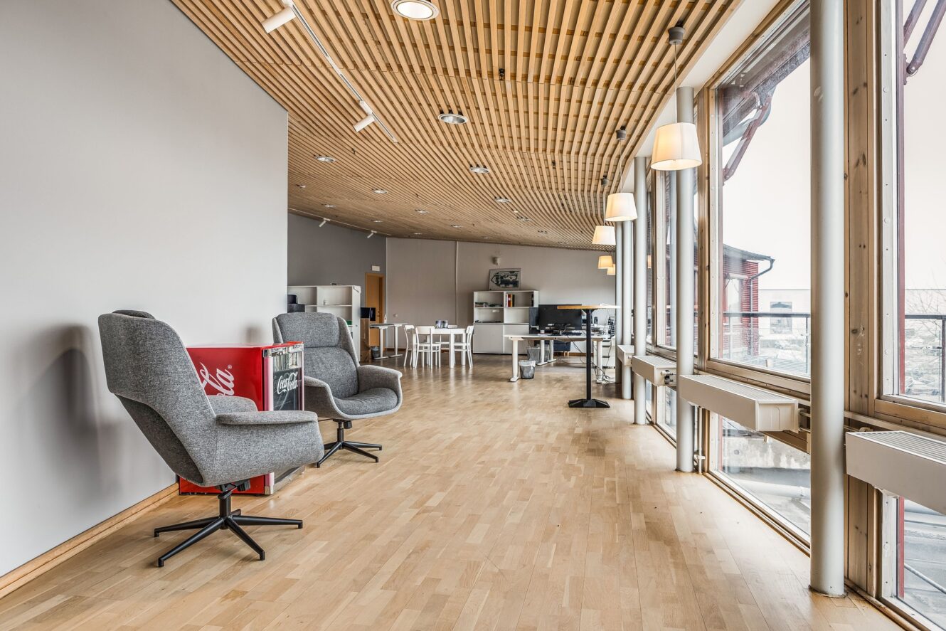 Modernt loungeområde med trägolv och tak, stora fönster, grå fåtöljer, en röd varuautomat och bord med stolar i bakgrunden.