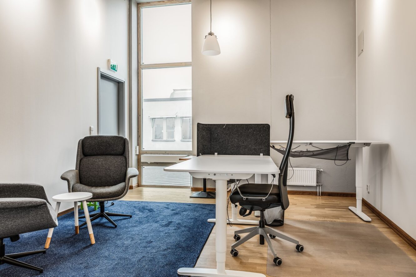 Modernt kontorsrum med blå matta, två fåtöljer, ett vitt runt bord och ett skrivbord med en kontorsstol nära ett stort fönster och vita väggar.