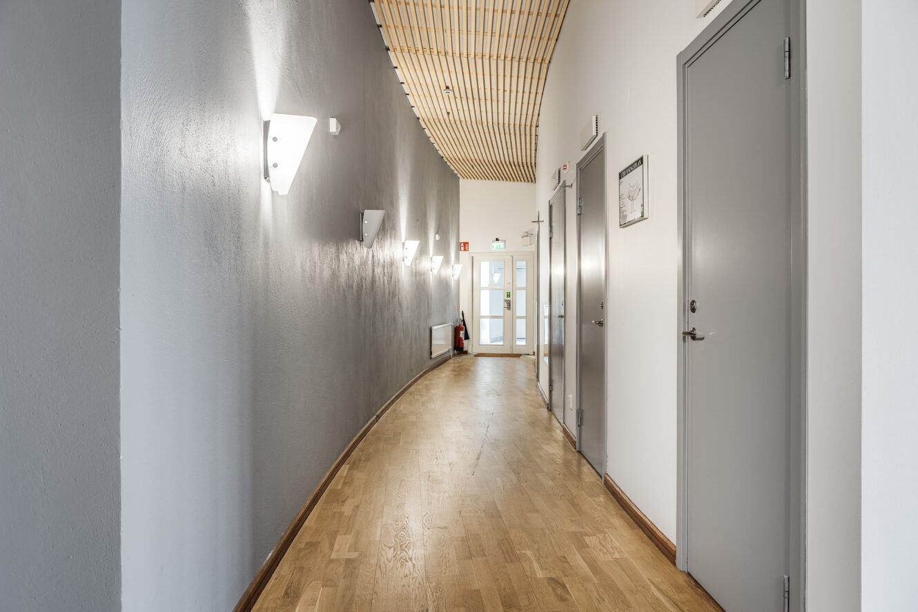 En modern hall med trägolv, svängda grå och vita väggar, grå dörrar till höger och väggmonterade lampor till vänster.