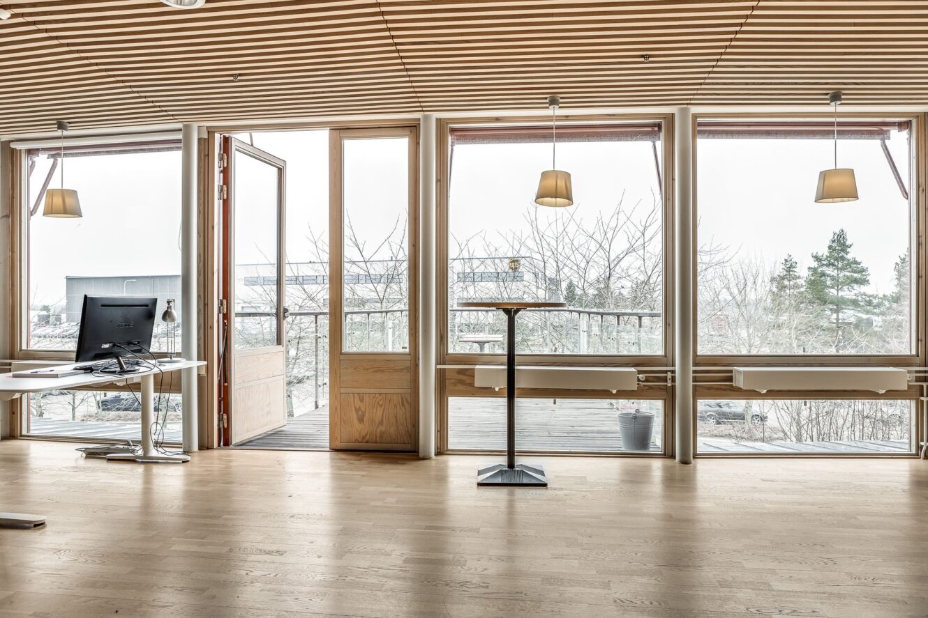 Modernt kontor med trägolv och tak, stora fönster, ett skrivbord med dator och en dörr som leder ut till en balkong med utomhusutsikt.