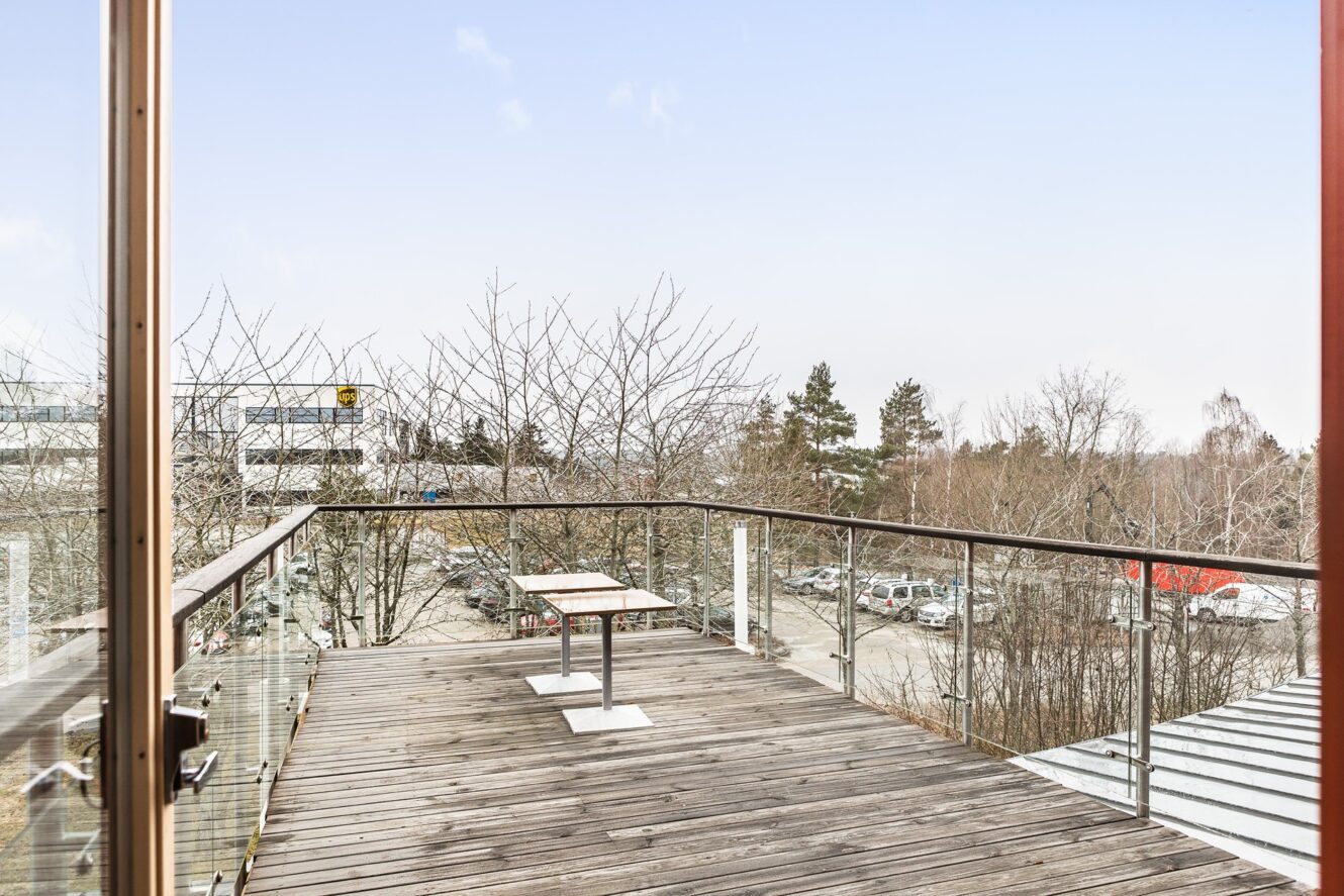 Från en träbalkong med glasräcken och två bord har man utsikt över en parkeringsplats och träd när det är klart väder.