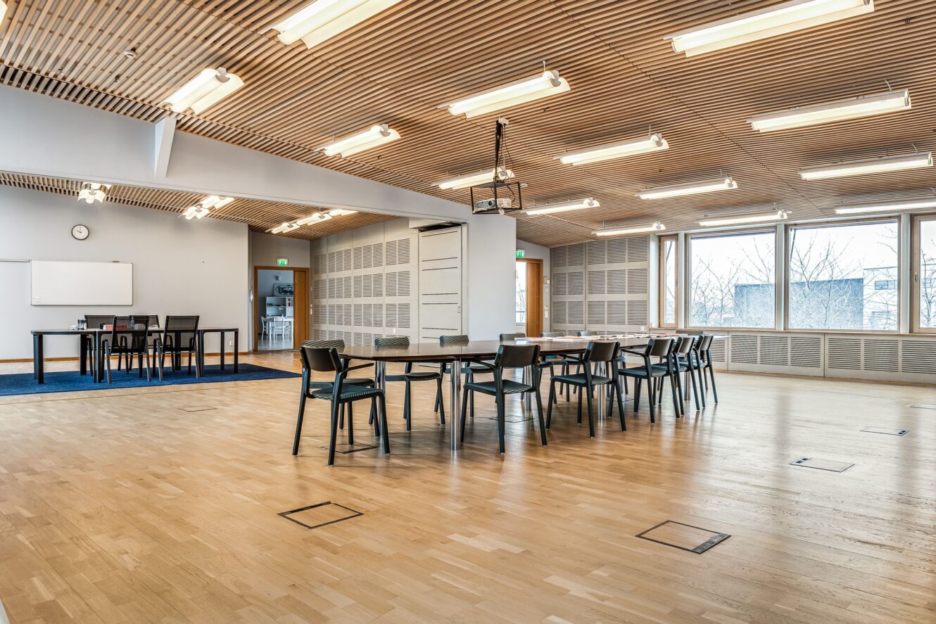 Rymligt, modernt klassrum med trägolv, flera bord och stolar, stora fönster, whiteboard, digital projektor och takbelysning.
