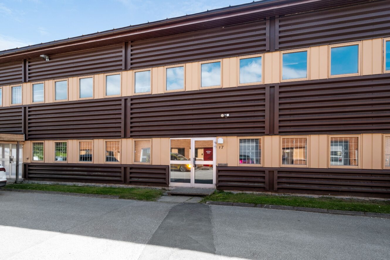 Tvåvåningsbyggnad med brun fasadbeklädnad, beige fönsterkarmar och en ingångsdörr i glas med siffran 17; bilar speglas i fönstren.