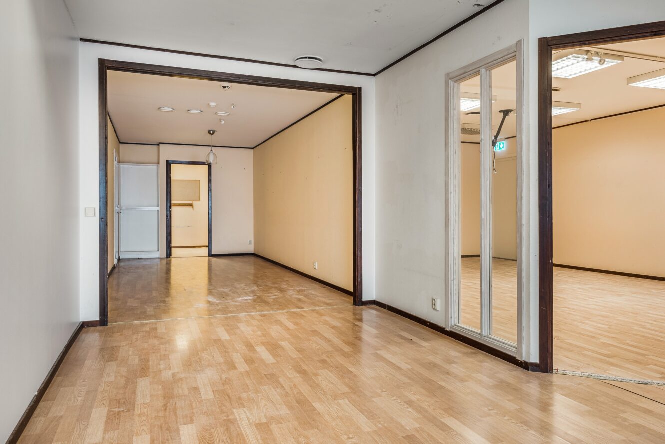 Tomt kontorsutrymme med ljust trägolv, beige väggar, lysrörsbelysning och flera dörröppningar som leder till angränsande rum.