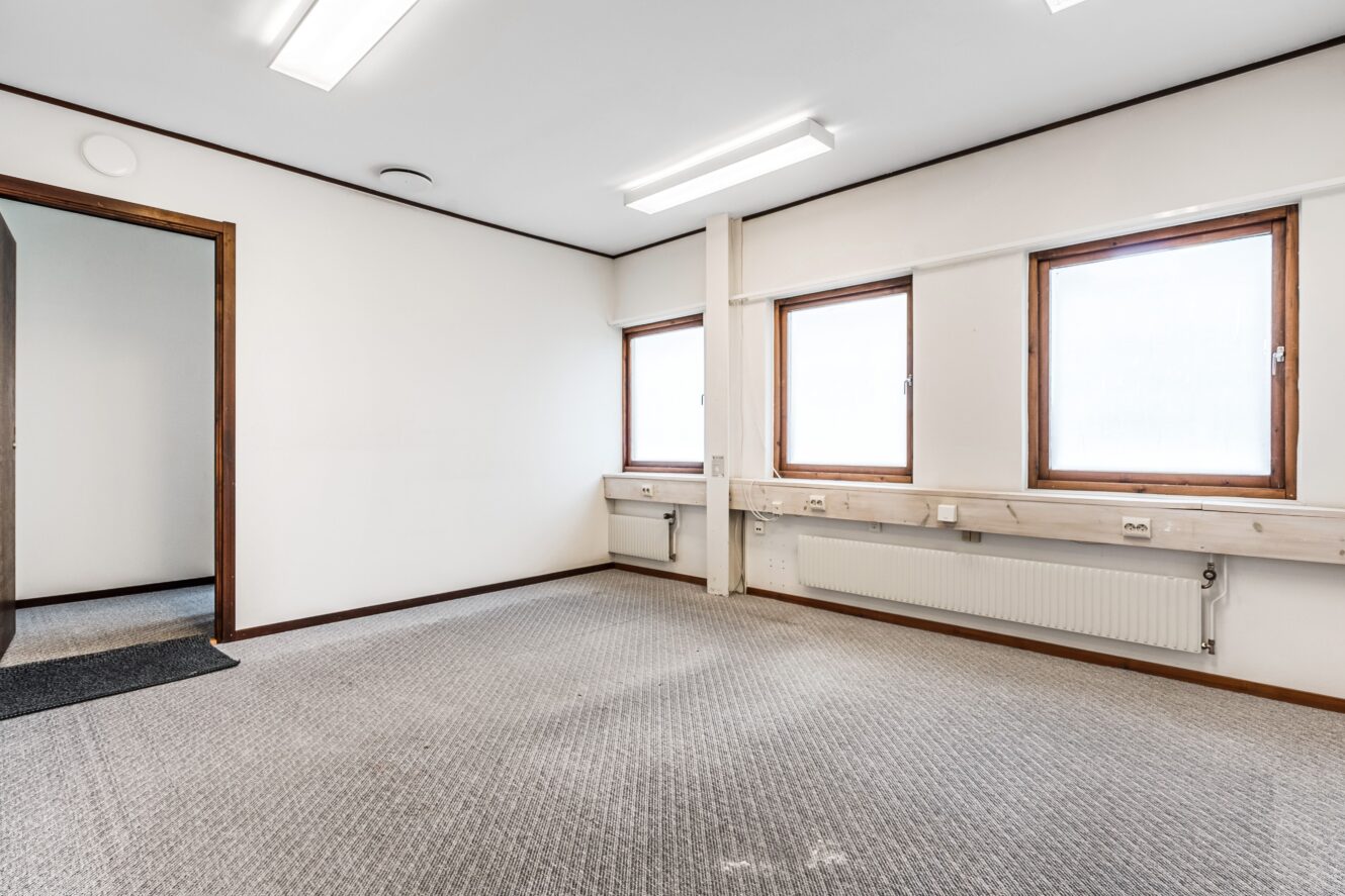 Tomt kontorsrum med tre fönster, vita väggar, grå heltäckningsmatta, lysrör i taket och synliga radiatorer och eluttag längs väggen.