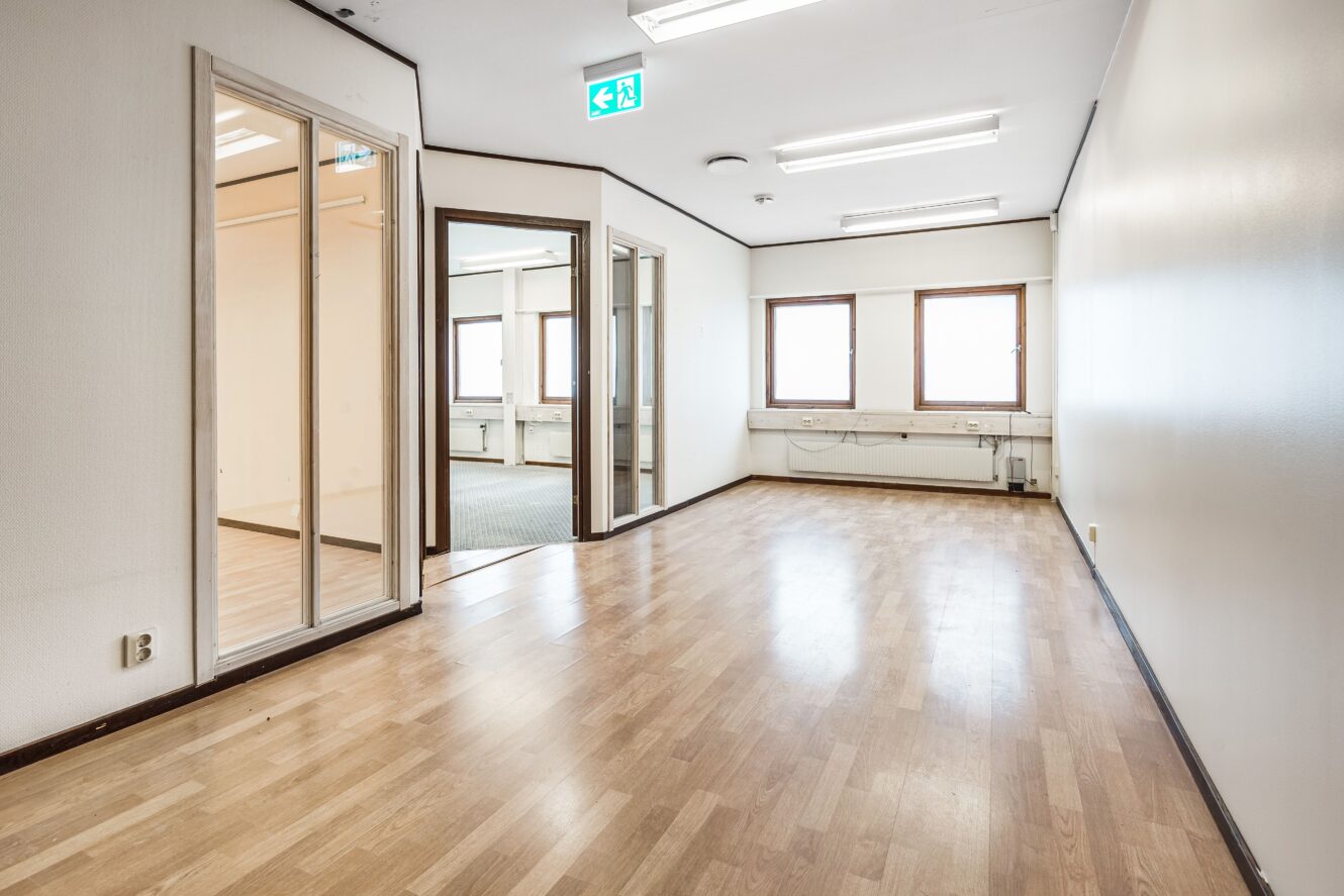 Tomt kontorsutrymme med ljust trägolv, vita väggar, tre fönster, lysrörsbelysning och rum med glasväggar.