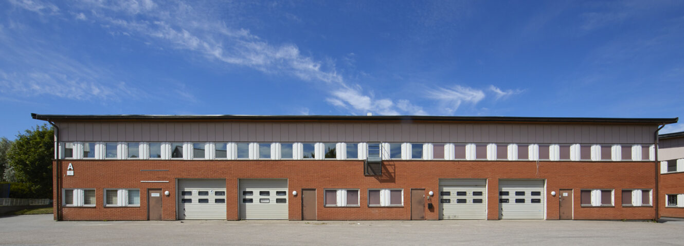 En tvåvånings industribyggnad med tegelfasad, flera garageportar och rektangulära fönster under en klarblå himmel.