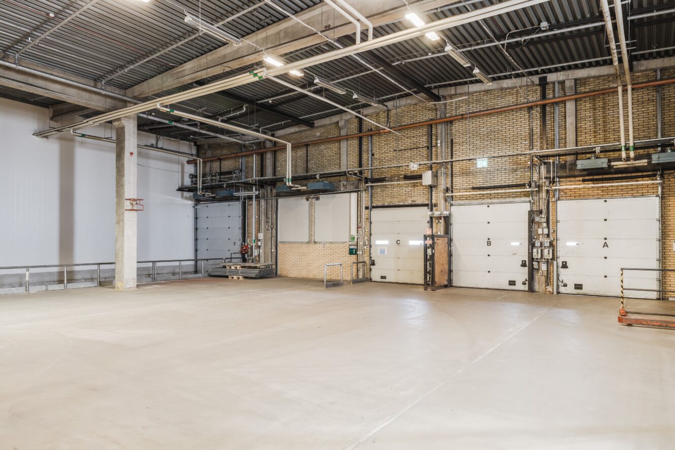 Rymlig, tom lagerlokal med betonggolv, högt i tak, synliga rör, tegelväggar och flera stora industriella garageportar.
