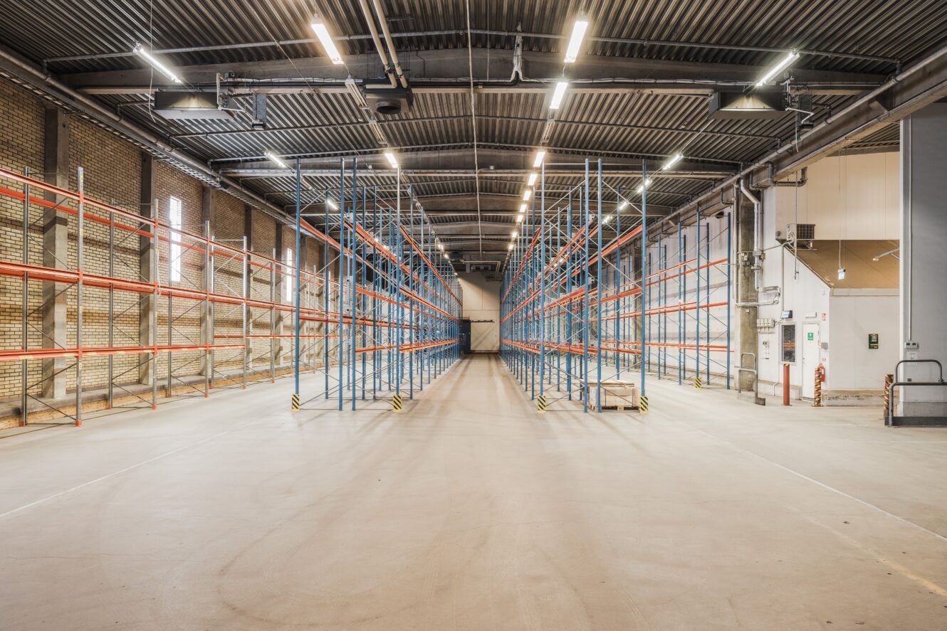 Tom lagerlokal med höga metallhyllor, högt i tak, synliga rör och stark takbelysning.