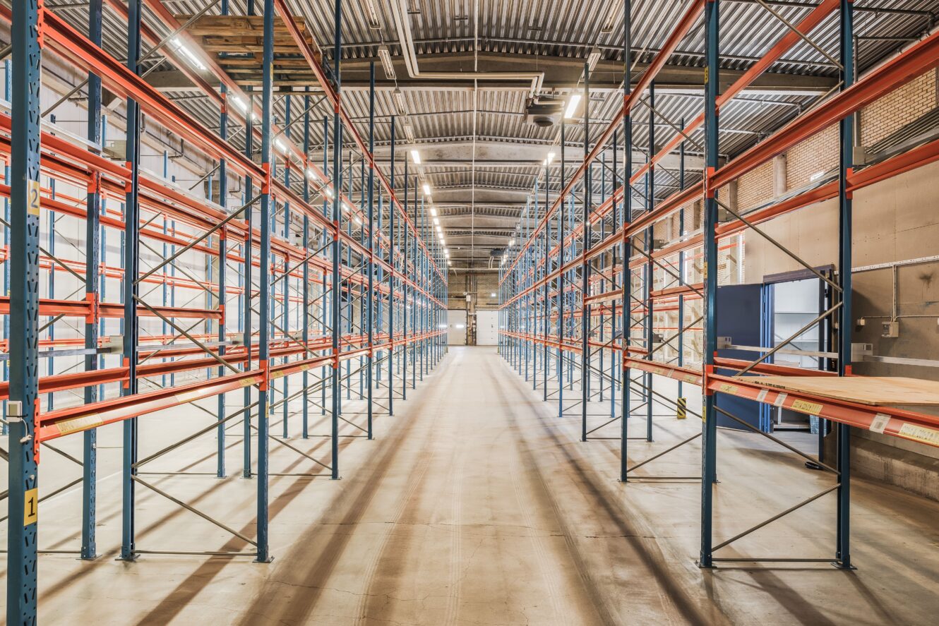 Tomt industrilager med lagerhyllor i metall, betonggolv och högt obehandlat tak. Hyllorna är placerade i rader och utrymmet är väl upplyst av taklampor.