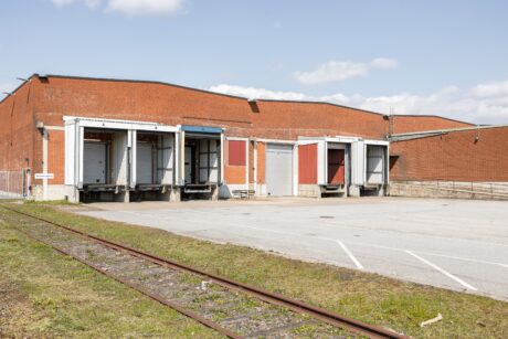 Lagerlokal i rött tegel med fyra lastkajer, flera öppna dörrar, tom parkeringsplats och ett nedlagt järnvägsspår i förgrunden under en klar himmel.