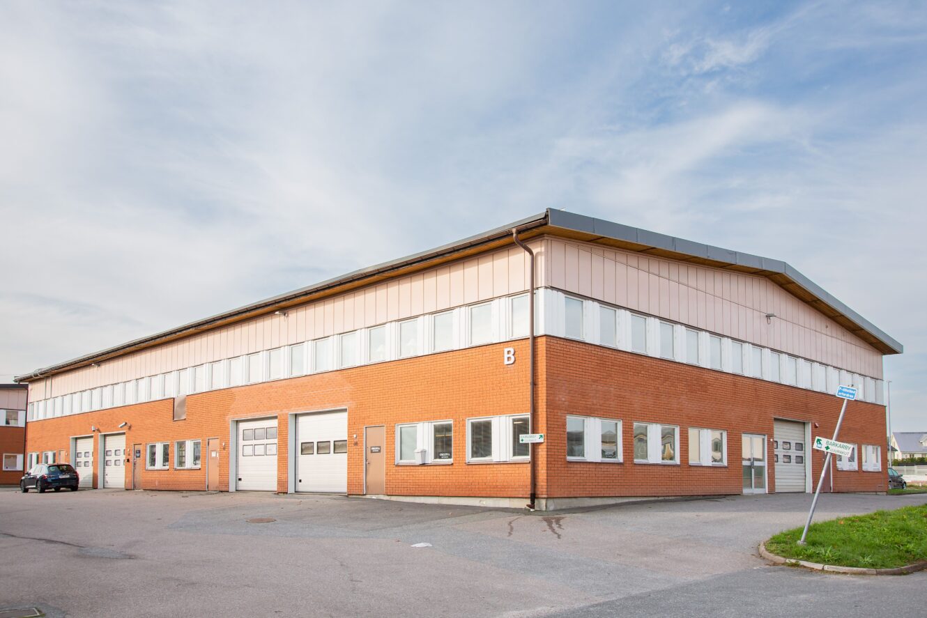 En stor rektangulär industribyggnad med underväggar i rött tegel, beige överväggar, flera garageportar och flera fönster, belägen på en asfalterad tomt under en delvis molnig himmel.