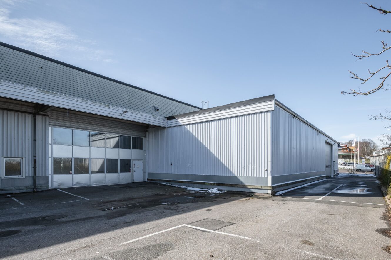 Ett stort, grått industrilager med metallfasad, en stor garageport och en tom parkeringsplats under en klarblå himmel.