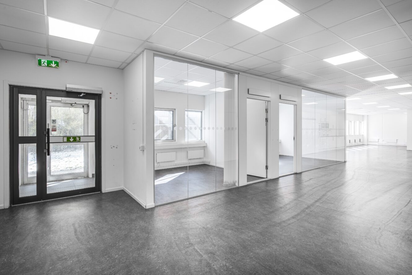 En modern kontorsinredning med glasväggar, vita väggar, taklampor och mörkt golv. En nödutgångsdörr syns till vänster.