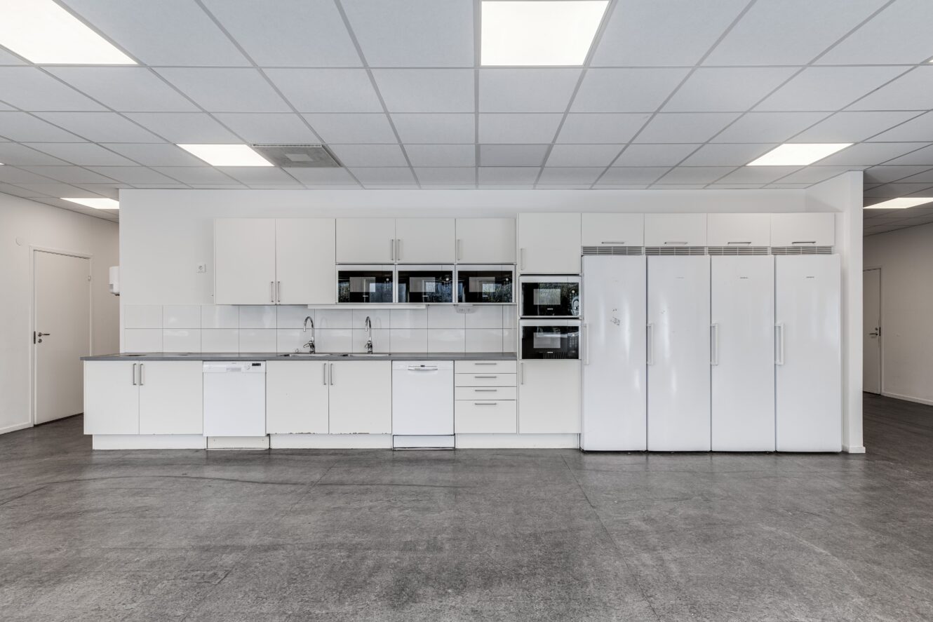 Modernt, minimalistiskt kök med vita skåp, flera vitvaror, inklusive ugnar och kylskåp, och ett grått golv under ett rutmönstrat tak med lysrörsbelysning.