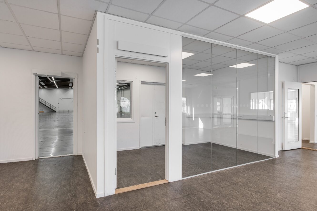 Modernt tomt kontor med mötesrum med glasväggar, vita väggar och synliga intilliggande lagerlokaler genom öppna dörröppningar.