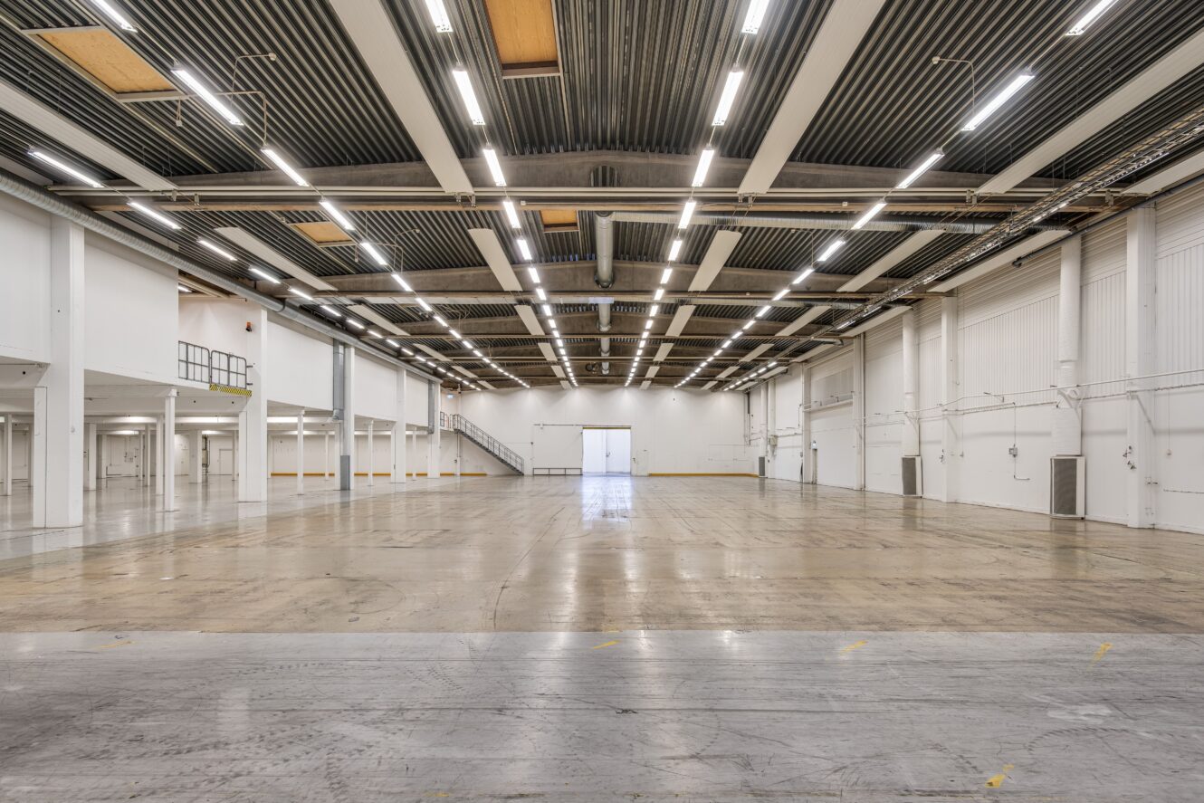 Stor tom industrilokal med högt i tak, synlig belysning och polerade betonggolv, sedd från ena änden mot en ljus utgång på andra sidan.