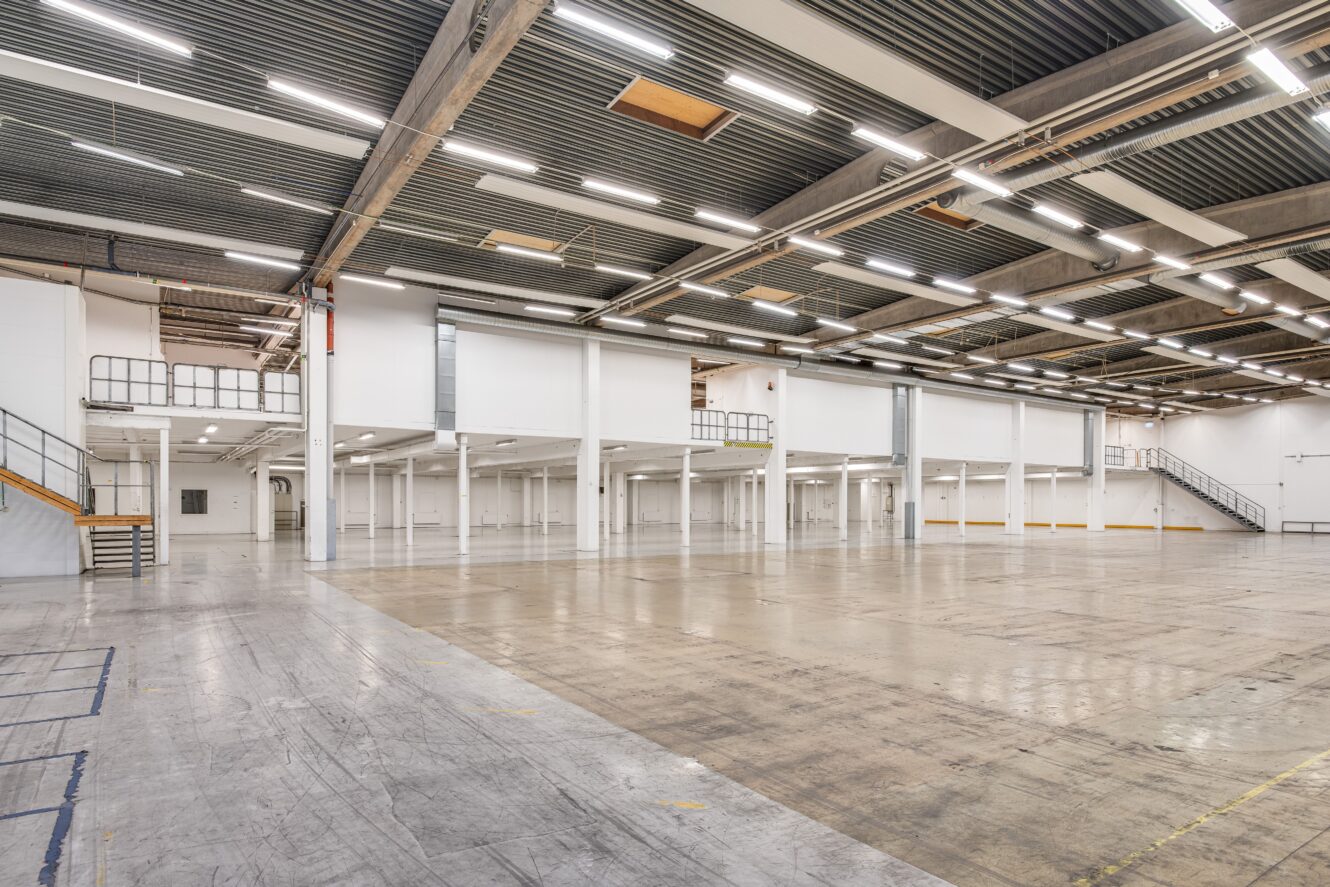 Rymlig, tom lagerlokal med högt i tak, synliga bjälkar, lysrörsbelysning och mezzaninvåningar längs väggarna.