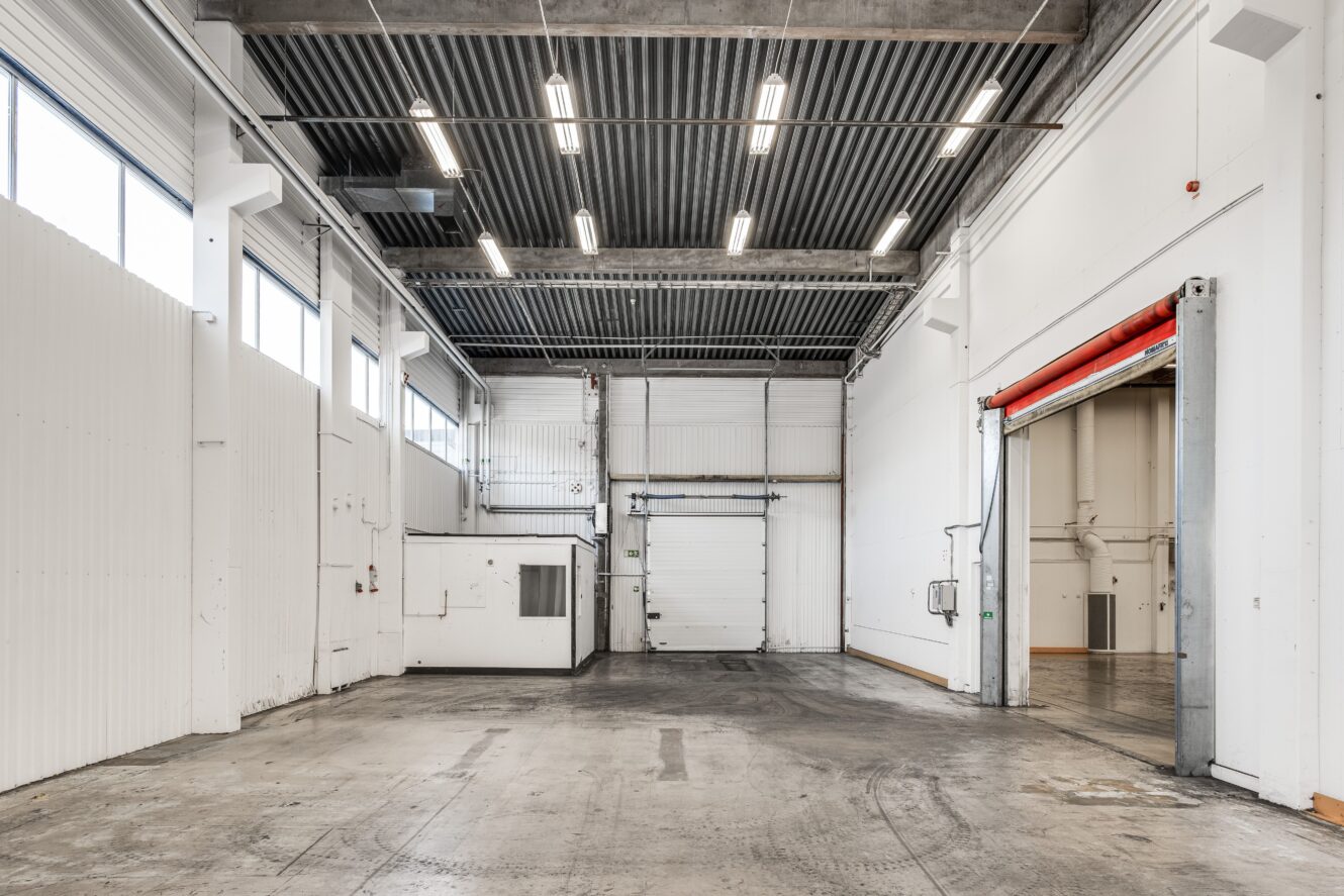 Tomt industriellt lagerutrymme med högt i tak, betonggolv, takbelysning, stora garageportar och vita väggar.