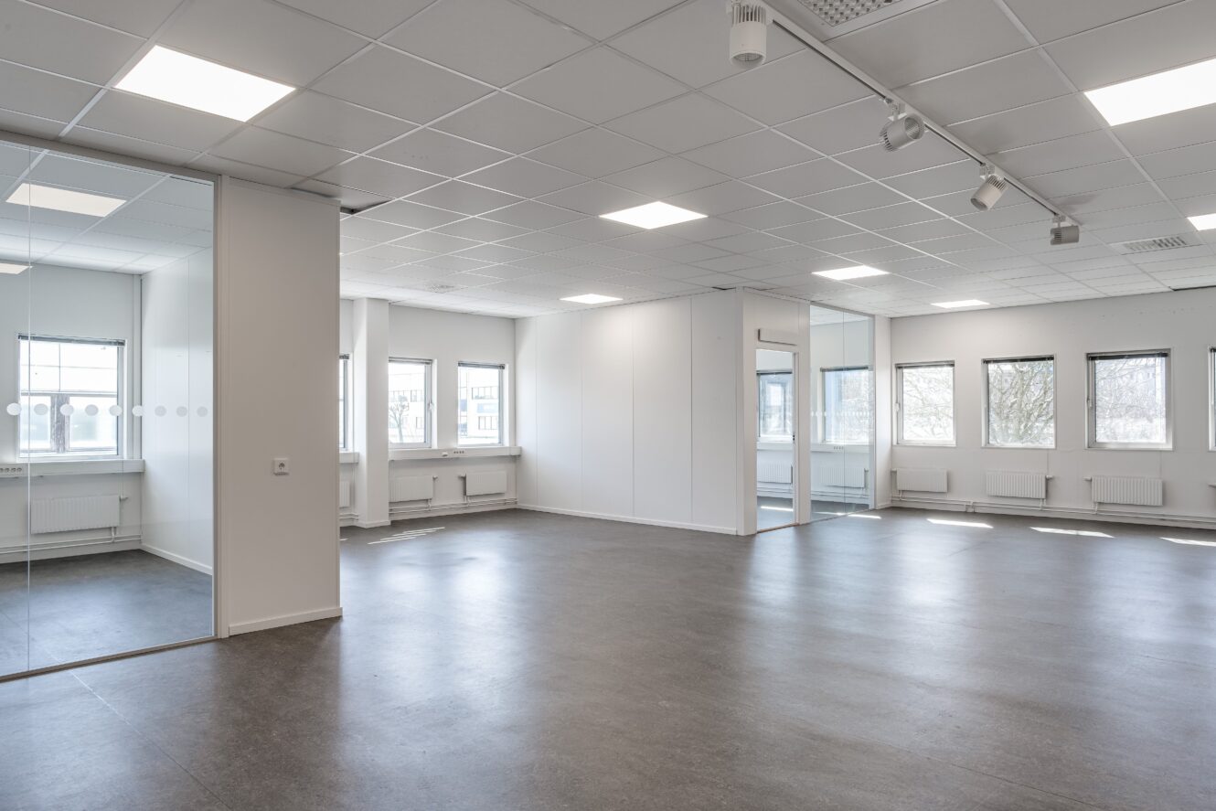 Tomt modernt kontorsutrymme med vita väggar, stora fönster, glasvägg och grått golv; taklampor och naturligt ljus lyser upp rummet.