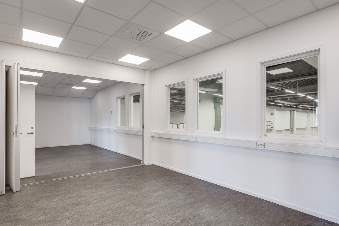 Tomt, modernt kontorsutrymme med vita väggar, tre innerfönster, öppna dubbeldörrar, grått golv och taklampor.
