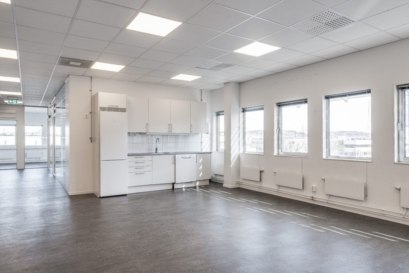 En tom, modern kontorslokal med vita väggar, ett litet pentry, stora fönster och grått golv.