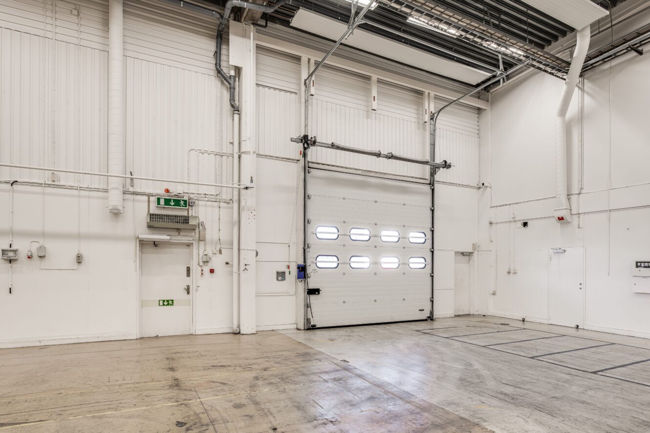 Tom industrilagerinredning med högt i tak, vita väggar, takbelysning och en stor stängd rulljalusidörr med små fönster.