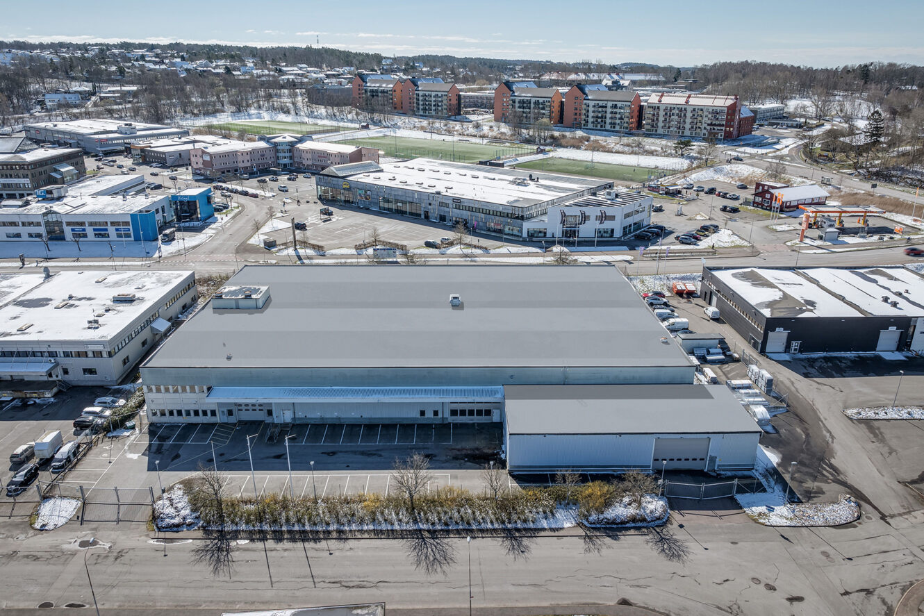 En stor industribyggnad med ett platt grått tak och en intilliggande parkeringsplats, omgiven av andra kommersiella byggnader och bostadshus en klar dag.