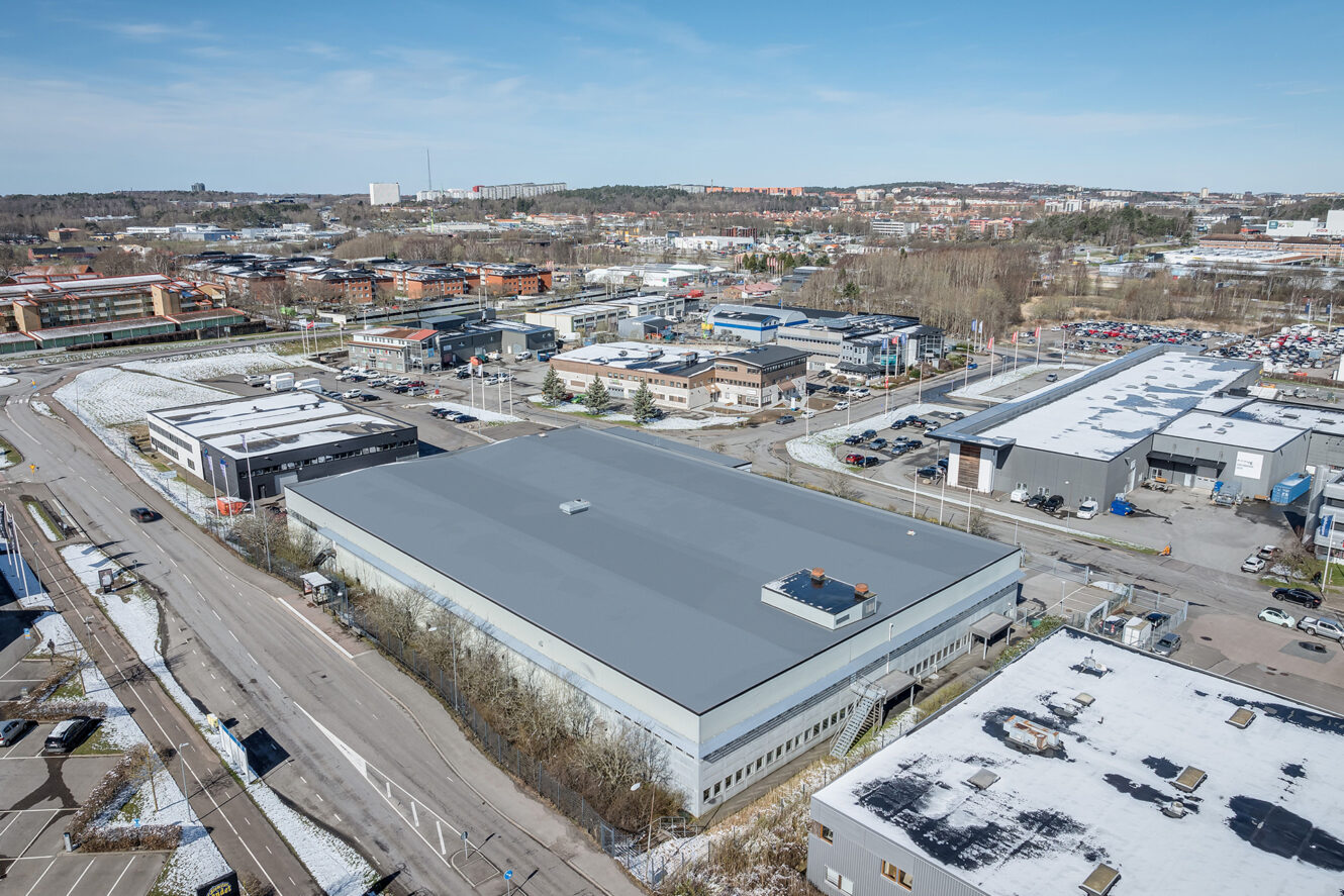 Flygfoto över ett industriområde med flera stora lagerbyggnader, parkeringsplatser och omgivande vägar under en klar himmel.