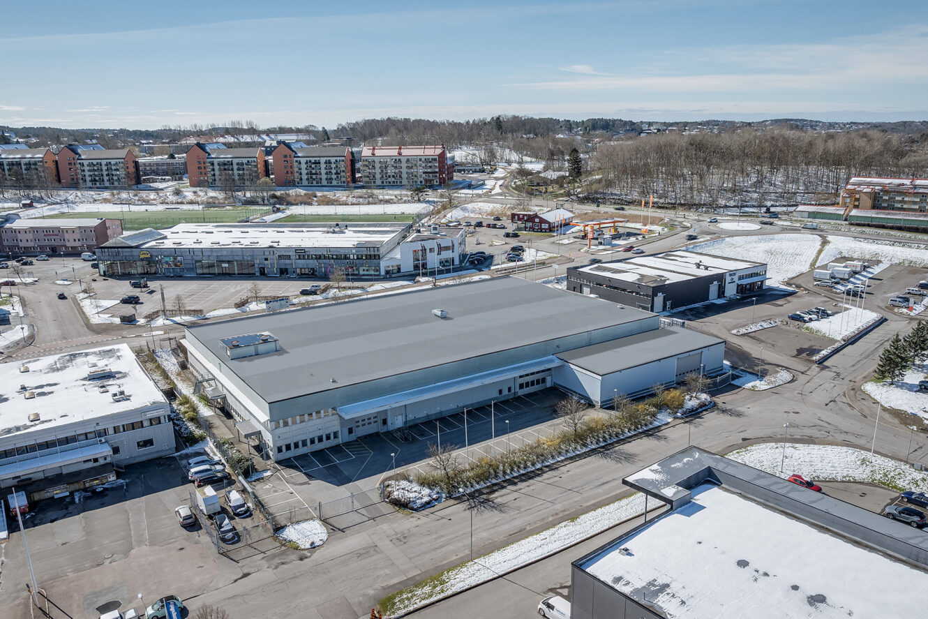 Flygfoto över en stor industribyggnad med platt tak omgiven av parkeringsplatser, andra kommersiella byggnader och snöfläckar. Bostadshus syns i bakgrunden.