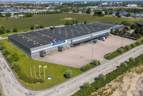 Flygfoto över ett stort industrilager med lastkajer, omgivet av grönska och parkeringsplatser, beläget nära fält och andra kommersiella byggnader.