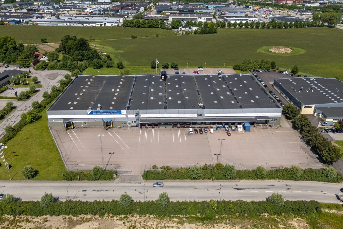 Flygfoto över ett stort industrilager med parkeringsplats, lastkajer och flera parkerade fordon, omgivet av vägar, grönska och andra byggnader.