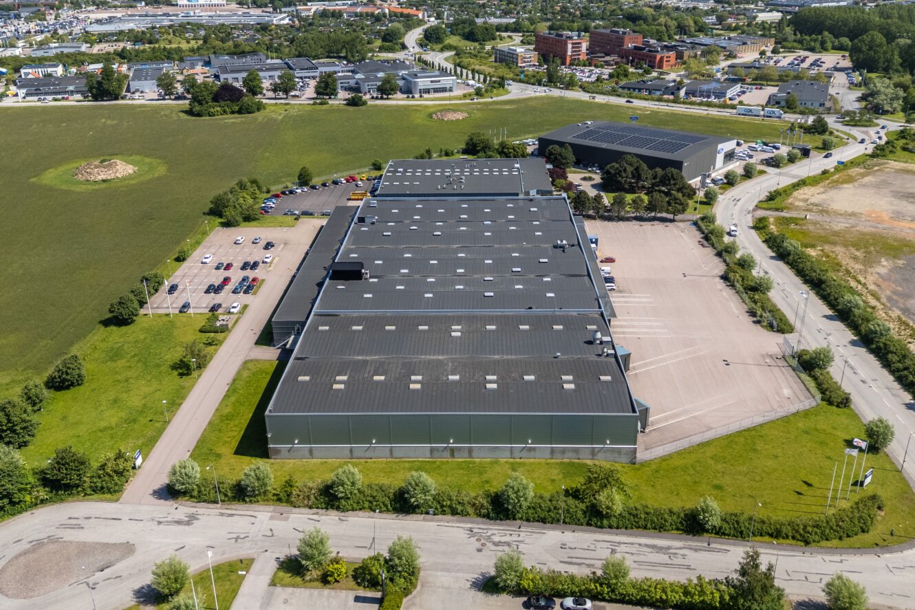 Flygfoto över en stor industrihall med mörkt tak, omgiven av parkeringsplatser, vägar och grönområden i ett affärsdistrikt.