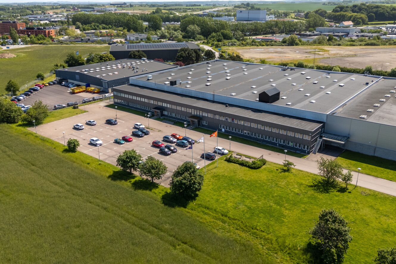 Flygfoto över en stor industribyggnad med en parkeringsplats, omgiven av gräs och träd, med flera bilar parkerade utanför.
