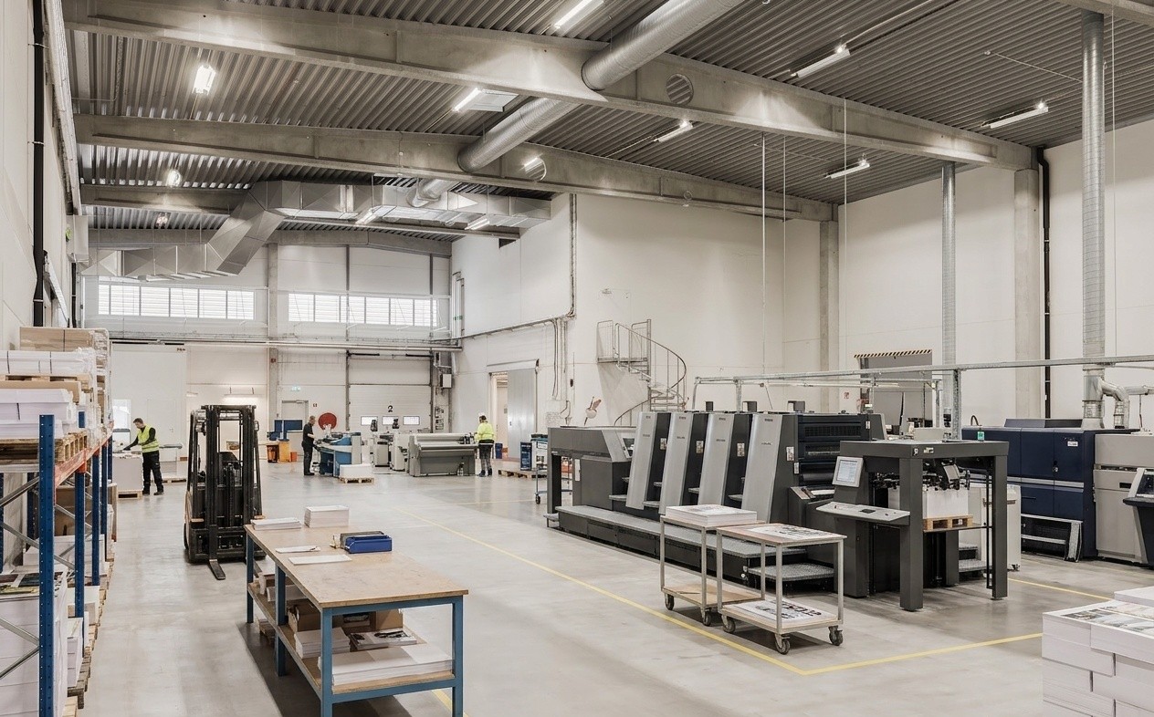 En stor, modern tryckanläggning med industriella tryckpressar, arbetsbord, pappersstaplar och arbetare i säkerhetsvästar i en väl upplyst lokal.