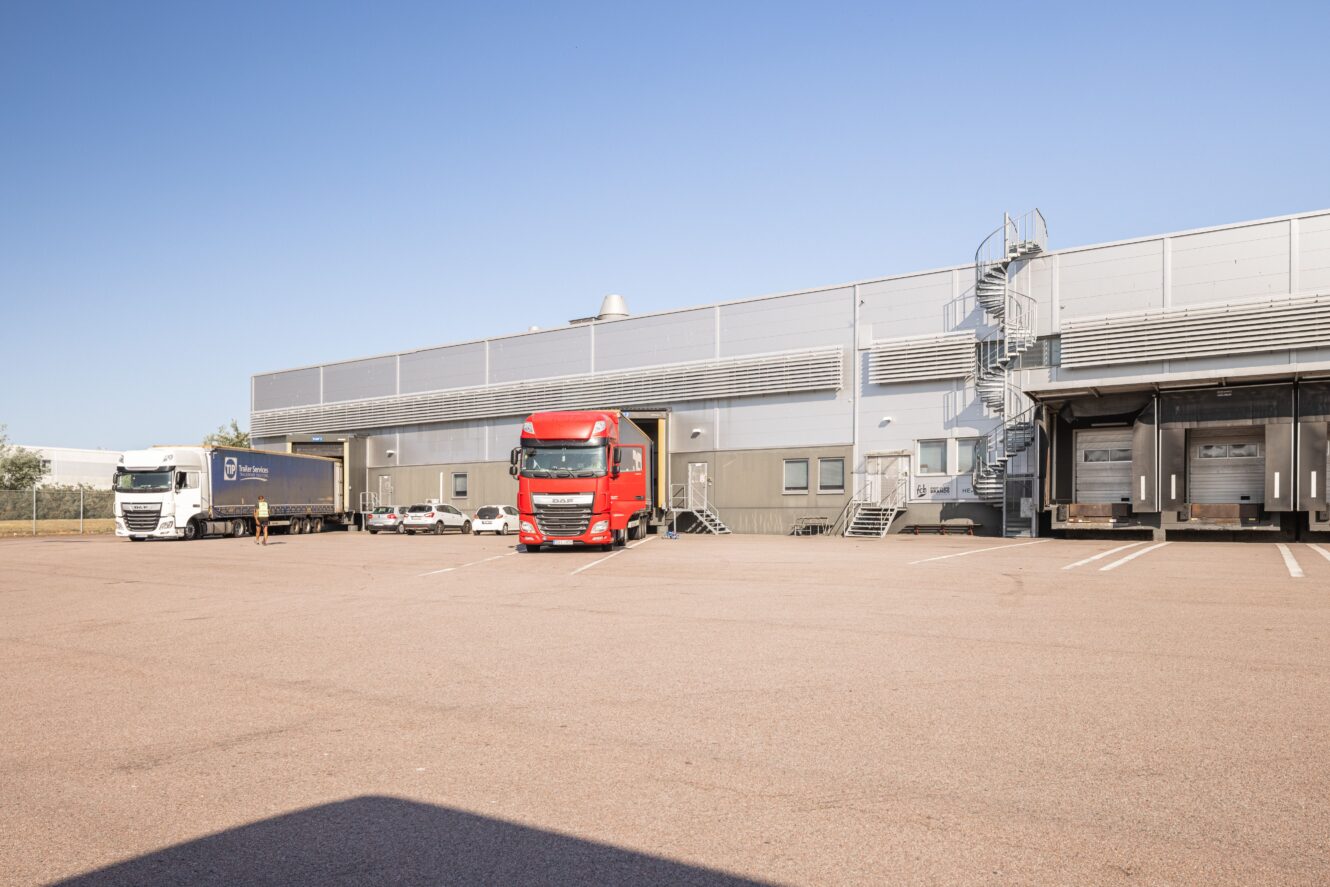 Tre lastbilar står parkerade utanför en modern lagerbyggnad med lastbryggor och en utvändig metalltrappa en klar, solig dag.