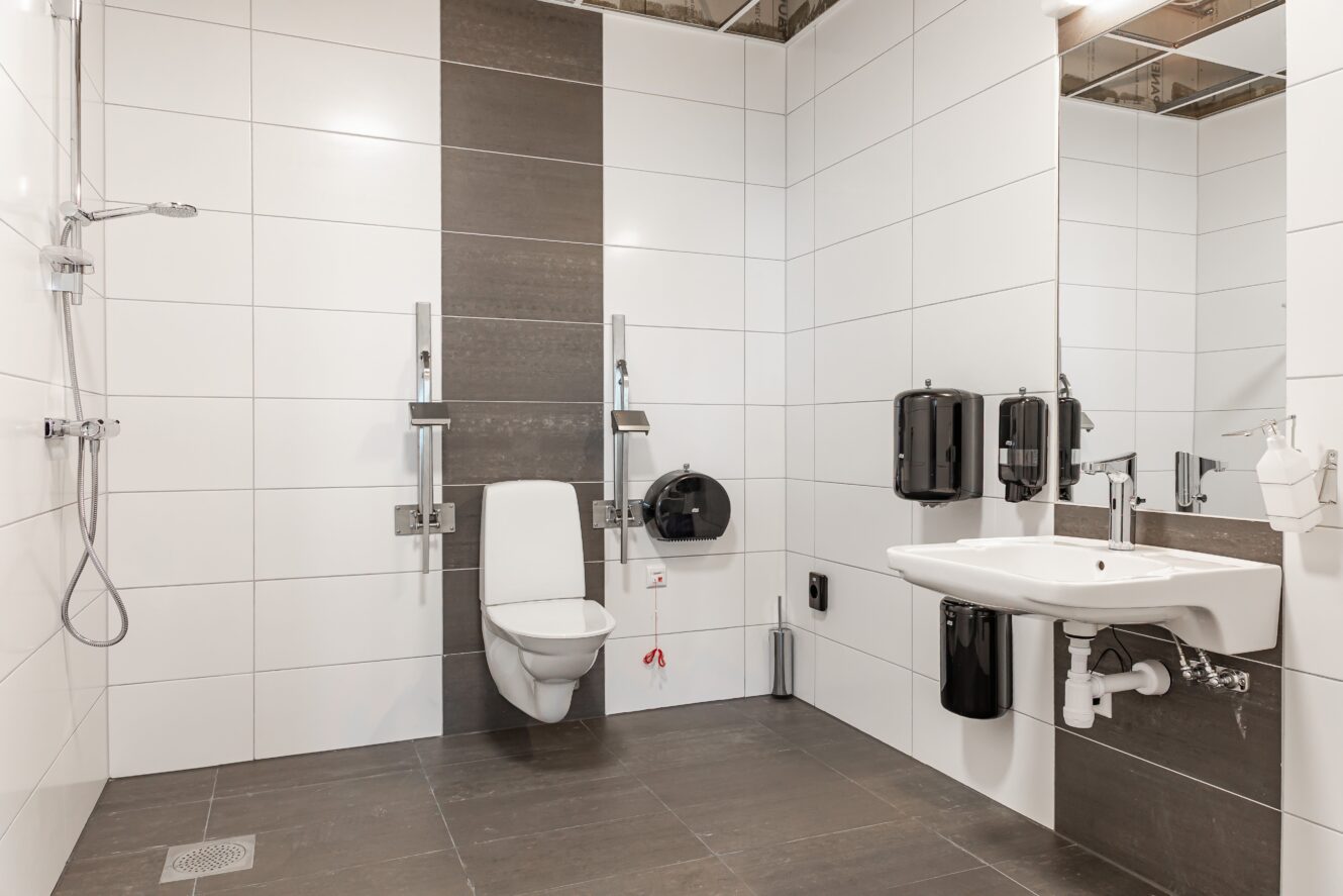 Tillgänglig offentlig toalett med vägghängd toalett, stödhandtag, handfat, tvål- och pappershandduksbehållare samt duschutrymme. Rummet har vitkaklade väggar och mörkgrå golvplattor.